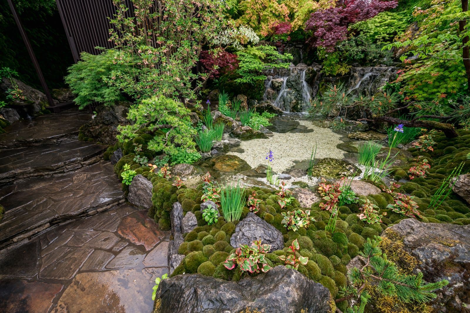A stone-paved path and a mossy waterside, with a waterfall beyond